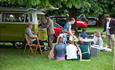 Family sat around in vintage clothing enjoying a nice picnic outside their green camper van