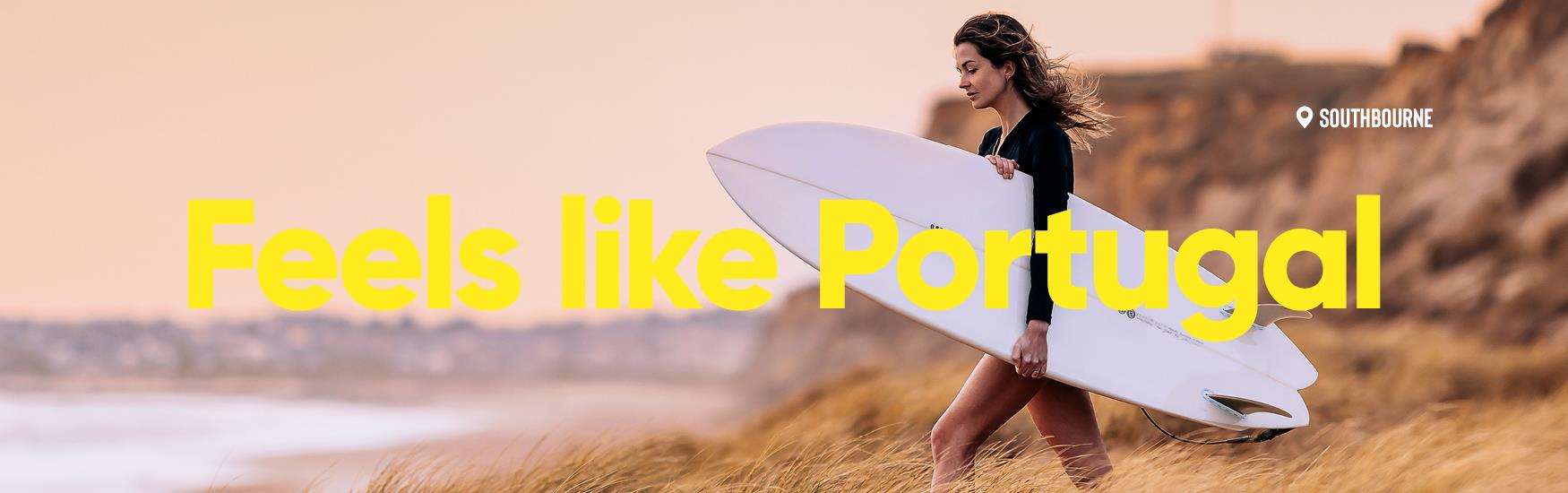 Woman walks with her surf board amogst the sand and grass towards the sea, text overlay reads "feels like Portugal"