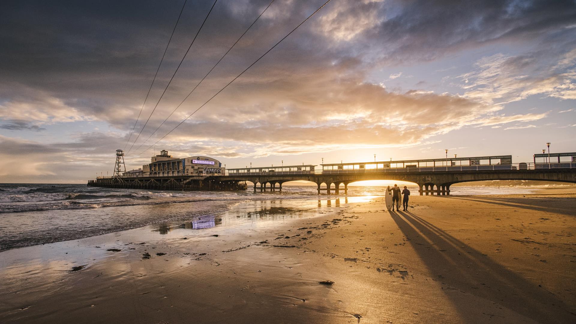 Two surfers walking on the beach at sunset with Bournemouth pier in the background