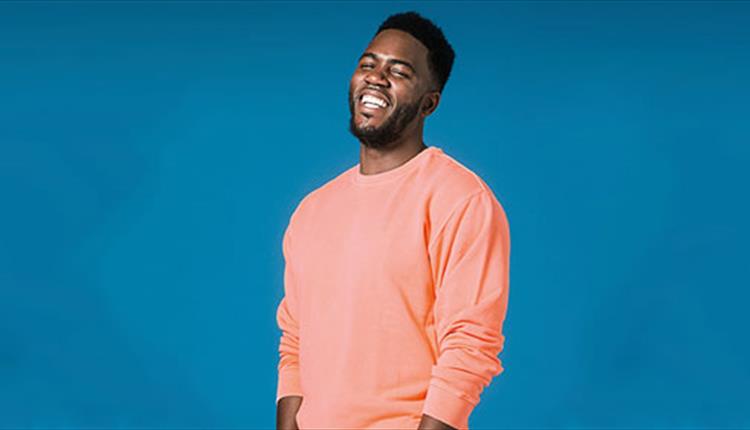 Comedian Mo Gilligan stands in a pink sweatshirt in front of a blue background.