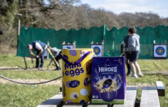 Two easter eggs on a bench in front of children doing archery.