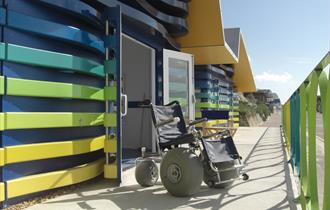 Beach wheelchait sat outside the green, blue and yellow accessible huts