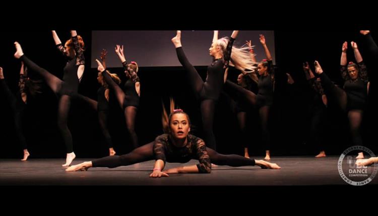 Dancers dressed in black leotards kick their legs above their heads. Principal dancer lies in splits, centre stage.