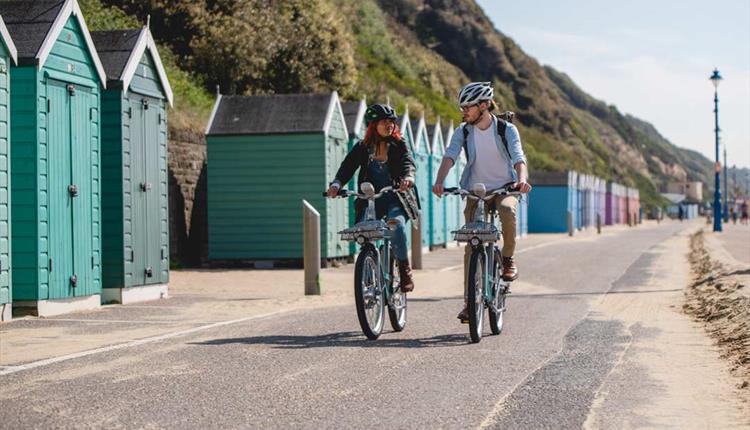 Couple cycling along Bournemouth seafront with beach huts behind them