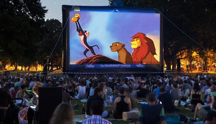 Large outdoor screen showing movie with people sitting on grass watching in the evening
