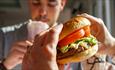 Close-up of hands holding a burger with a man blurred in the background drinking a milkshake