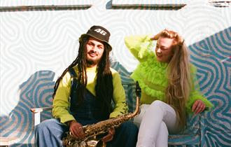 Blue and white swirly background with a man with dreadlocks and a woman in a brilliant green feathery top sitting. Man is holding a saxophone  and loo