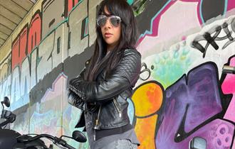 Lady posed in front of abstract artwork wall wearing leather jacket with arms crossed and wearing sunglasses
