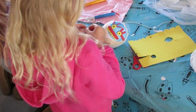 The image is of the back of the head of a blonde-haired child wearing a pink jumper, making a Halloween decoration at a table with a blue table cover