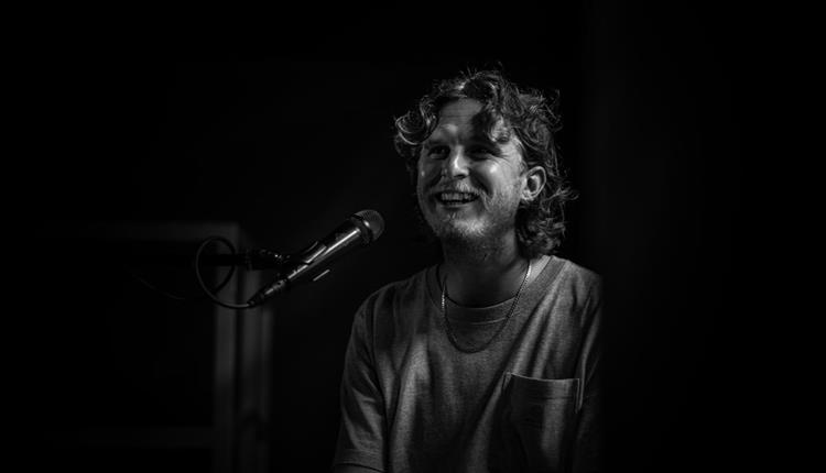 Black & white image of long curly haired man sitting in front of a microphone, smiling