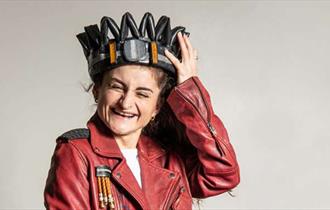 A woman with a red leather jacket and white top on, holding a crown on top of her head whilst giggling.