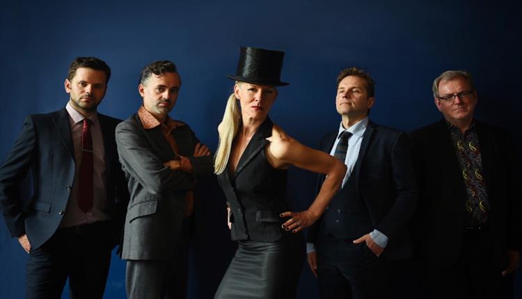 4 men stand against a dark blue backdrop in dark suits and ties. A woman stands in the centre of them with a black top hat.