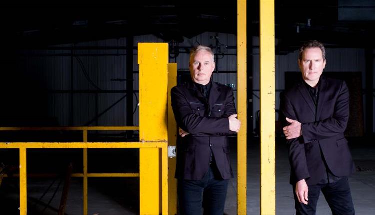 OMD Band Shot, 2 male band members, facing camera with serious expressions, wearing black suits against a dark background