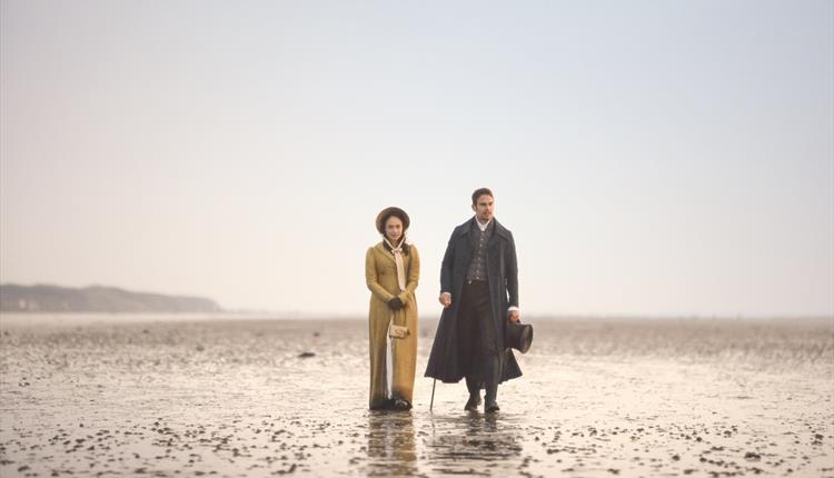 Rose Williams and Theo James walking along the beach with hazy sky