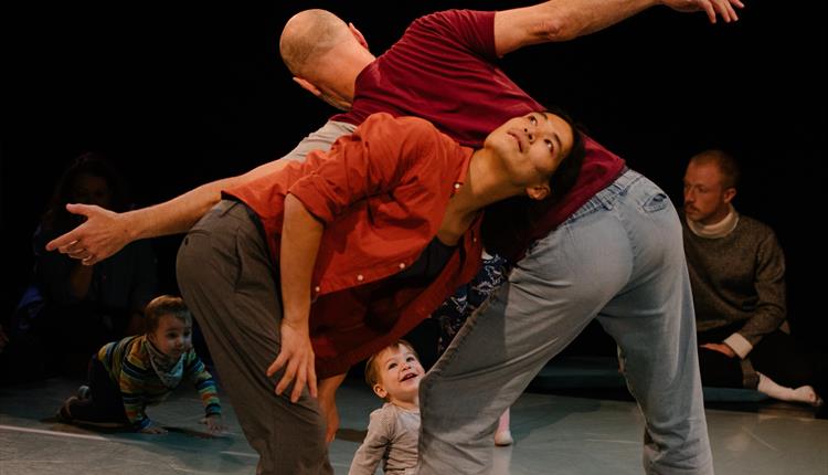 Two male dancers standing up making star shapes with a young child sitting down looking at them smiling.