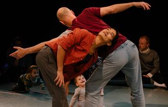 Two male dancers standing up making star shapes with a young child sitting down looking at them smiling.