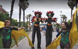 Stilt walkers in feathery costumes being led through the gardens by two ladies in green leotards with yellow capes