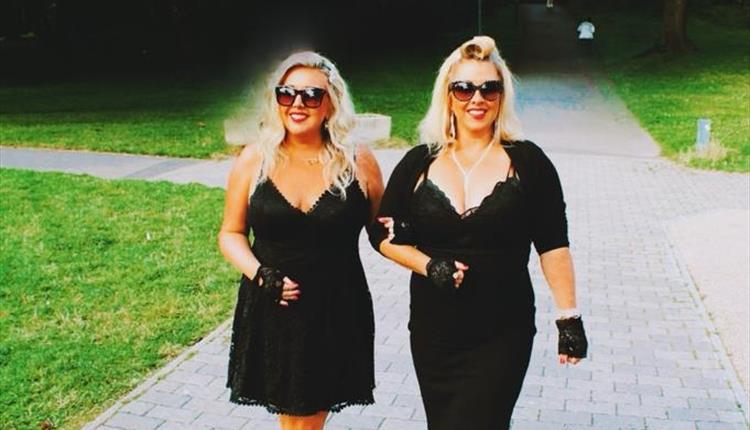 Two blond ladies in black dresses walking arm in arm