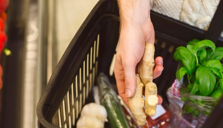 A supermarket basket with fresh produce and a hand holding a bulb of ginger