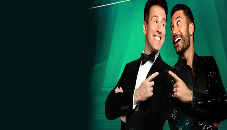 Anton Du Beke and Giovanni stand back to back smiling and pointing at each other. Green background.