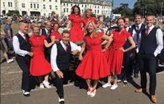 Dance troupe posed outside in front of buildings with ladies in red dresses and men in black waistcoats and white shirts - all facing front