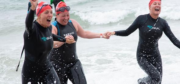 three women coming out the sea with wetsuits and swimming caps on their head laughing