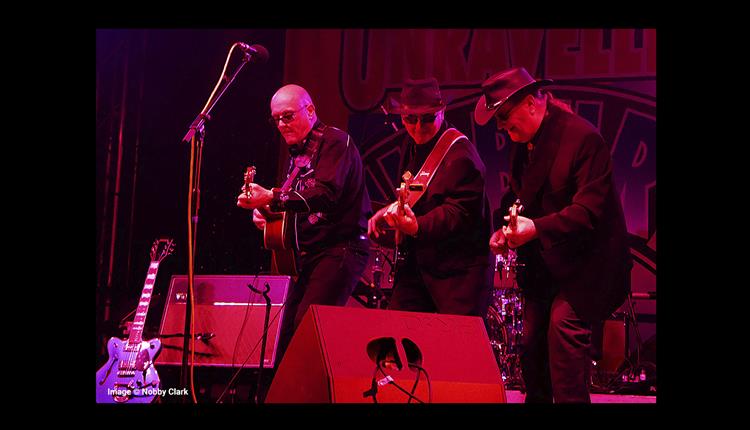 The Unravelling Wilburys on stage performing with guitars with a dark background