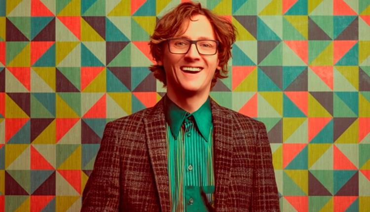 Ed Byrne smiling wearing a green shirt and checkered jacket with a bright background.