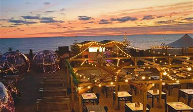 Image shows the Igloos at Brighton Music Hall, sparkling with fairy lights in front of a  beautiful Brighton sunset. 