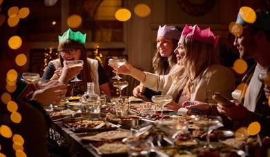 Partygoers enjoying a meal at Mowgli Brighton. a group of friends wearing colourful Christmas hats raise a toast, there are dishes of food in front of