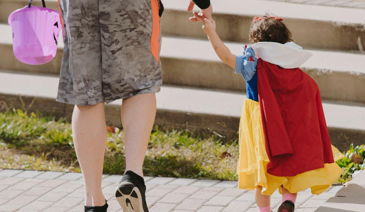 Little girl in costume with with an adult holding a purple pumpkin bucket