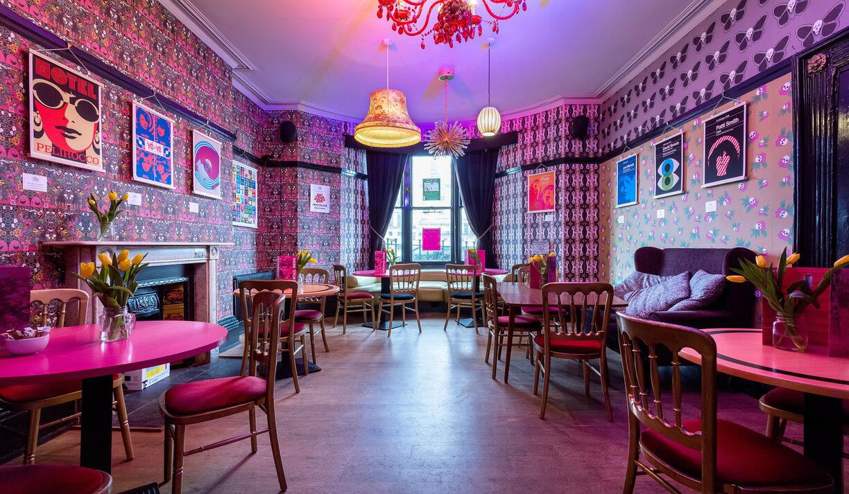 The bright, retro bar of the Hotel Pelirocco. The walls have pink patterned wallpaper and retro prints are hung around the space. The tables and chair