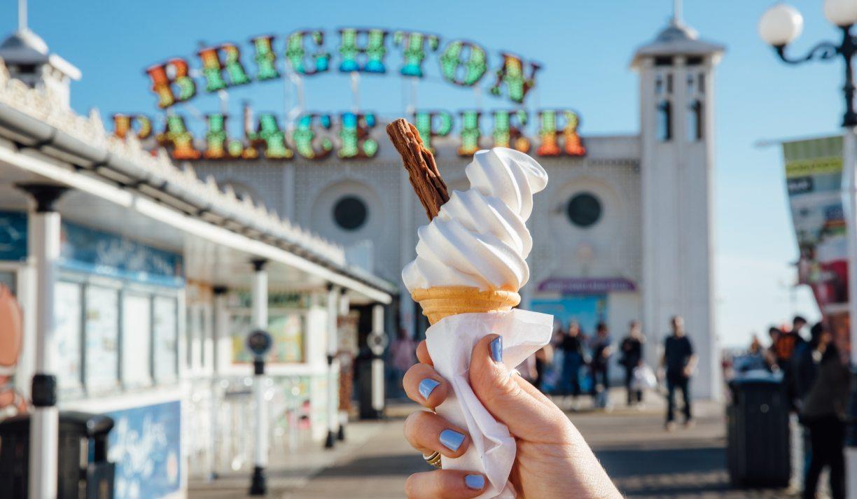 A hand holding an ice cream cone in front of the Brighton Palace Pier sign