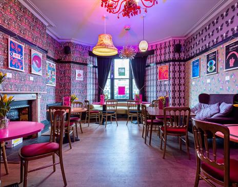 The bright, retro bar of the Hotel Pelirocco. The walls have pink patterned wallpaper and retro prints are hung around the space. The tables and chair