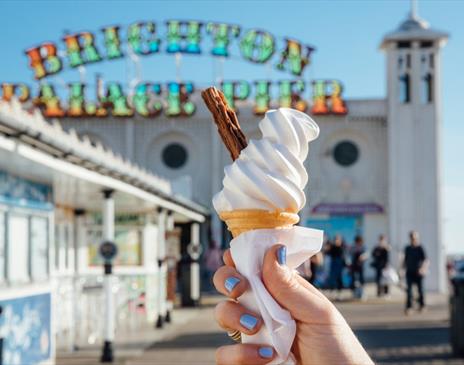 A hand holding an ice cream cone in front of the Brighton Palace Pier sign