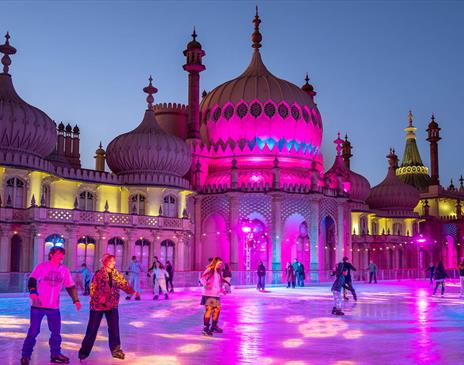 Image shows skaters at the Royal Pavilion ice rink, skating in front of the palace which is light in a warm pink glow.