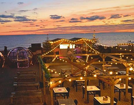 Image shows the Igloos at Brighton Music Hall, sparkling with fairy lights in front of a  beautiful Brighton sunset.