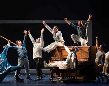 Actors on stage in Blizzard at Brighton Dome. Image shows actors in pale and blue outfits sitting on and around a piano on stage. They have their hand