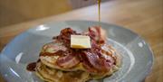 A plate of pancakes with bacon on top with a drizzle of maple syrup being poured from above.