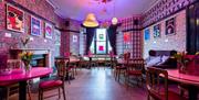 The bright, retro bar of the Hotel Pelirocco. The walls have pink patterned wallpaper and retro prints are hung around the space. The tables and chair