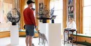 A man in a baseball cap and shorts is stood talking to a woman sat on a window seat. There is also a dog and plinths with sculptures on them. Photo cr