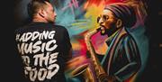 A painted mural on a wall of a playing a saxaphone next to a man wearing a T-Shirt that says #adding music to the food