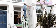 A person is up a ladder adding bunting to the outside of their house, underneath an Artists Open Houses banner. Photo credit: Syl Ojalla