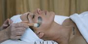 A person lying down on a bed with a hair wrap on, having a product applied to their face as part of a facial treatment.