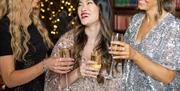 Image shows a group of festive party goers enjoying a glass of champagne while chatting, they are wearing sequined outfits.