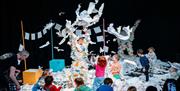 Image courtesy of Chloe Hashemi. Origami children's show at Brighton Dome Studio Theatre, children on stage with a performer throwing paper into the a