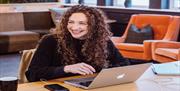 A person with long curly hair working at a laptop smiling