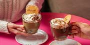 Two hands holding hot chocolates with cream and a wafer in them.