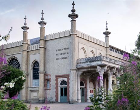 The image shows Brighton Museum & Art Gallery, a beautiful regency building with pink flowers in the foreground.