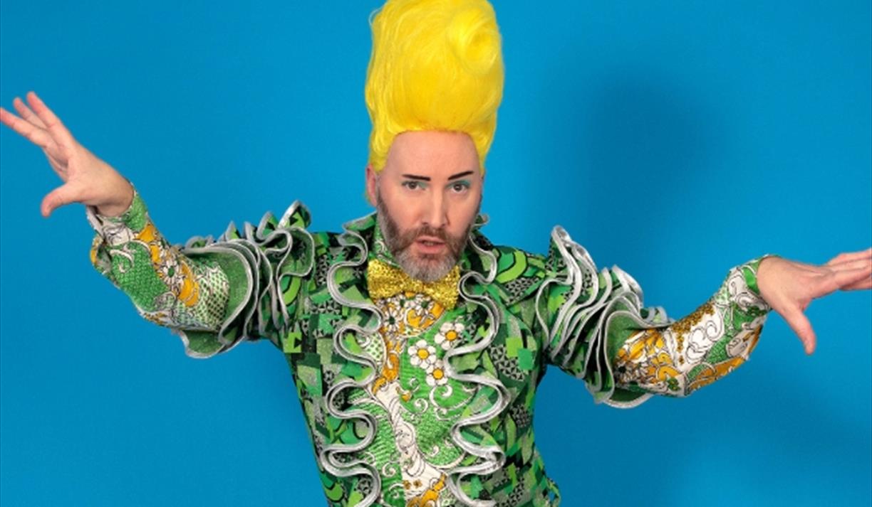 Image shows host Bugaloo Stu against a blue background wearing a yellow wig and green outfit with a ruffle on the front.